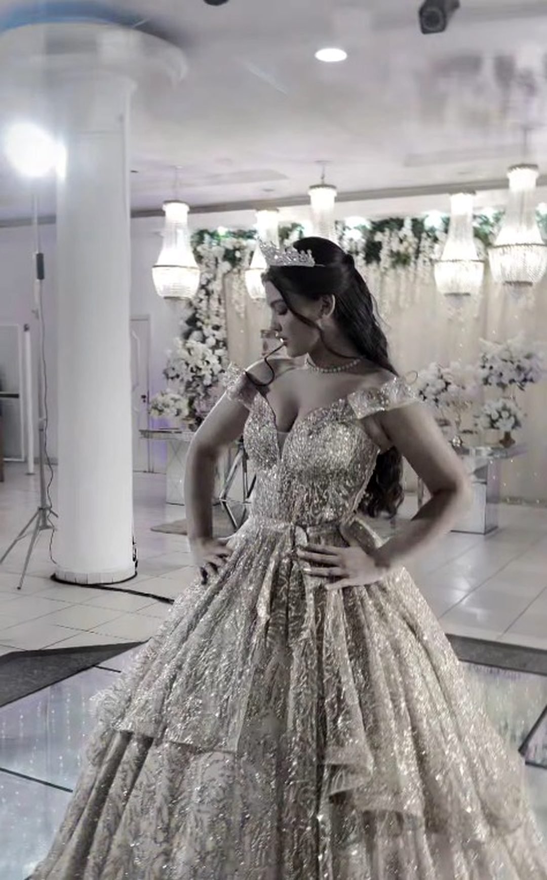 Debutante de coroa e vestido prateado no salão do Estrela Buffet, entre lustres e arranjos de flores