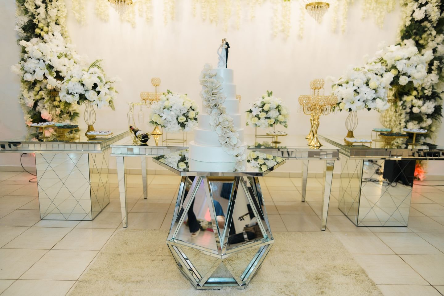 Mesa do bolo com base espelhada e arranjos de flores no salão do Estrela Buffet
