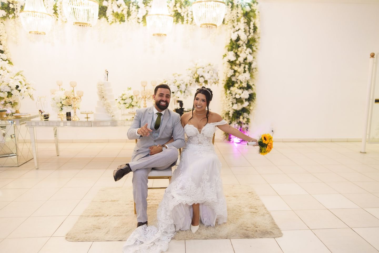 Noivos sorrindo no salão do Estrela Buffet, diante da mesa do bolo e do painel de flores