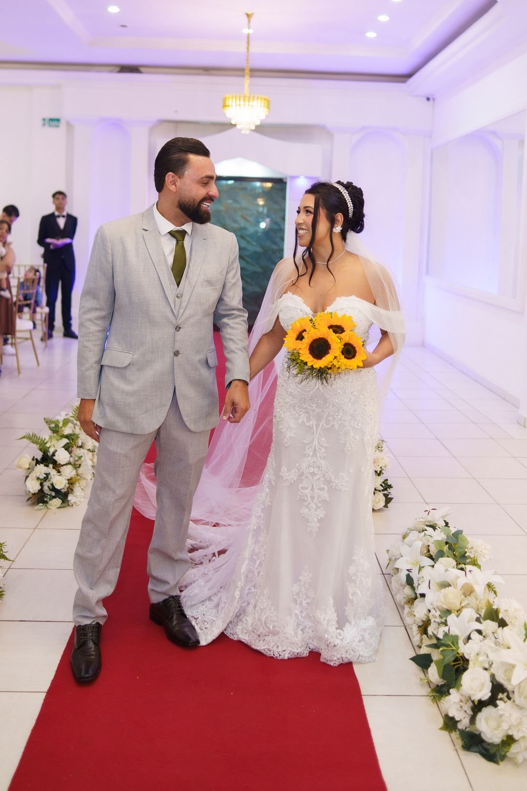 Noivos caminhando pelo corredor do salão do Estrela Buffet com buquê de girassóis