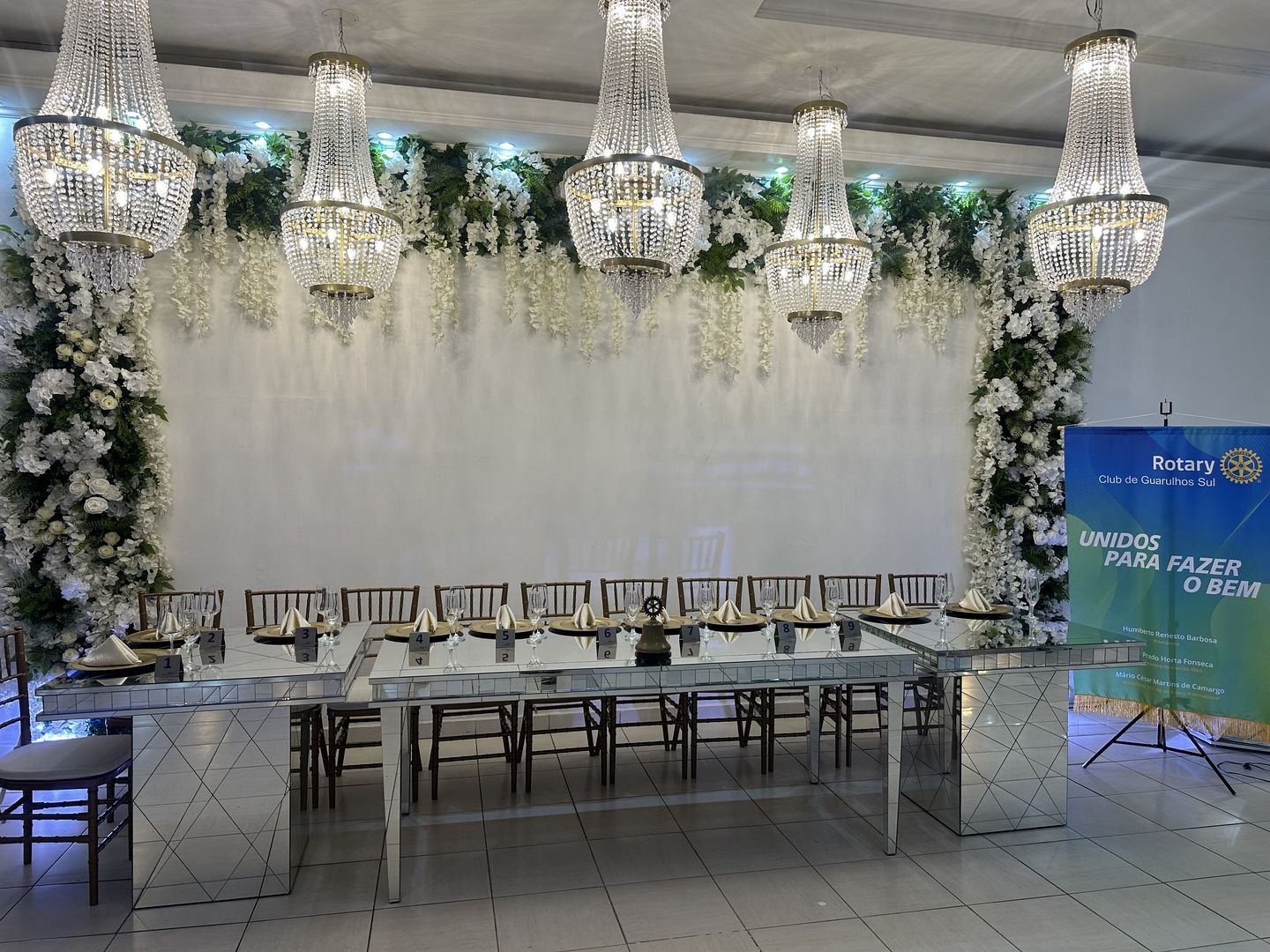 Salão montado com mesa imperial e painel de flores sob lustres de cristal