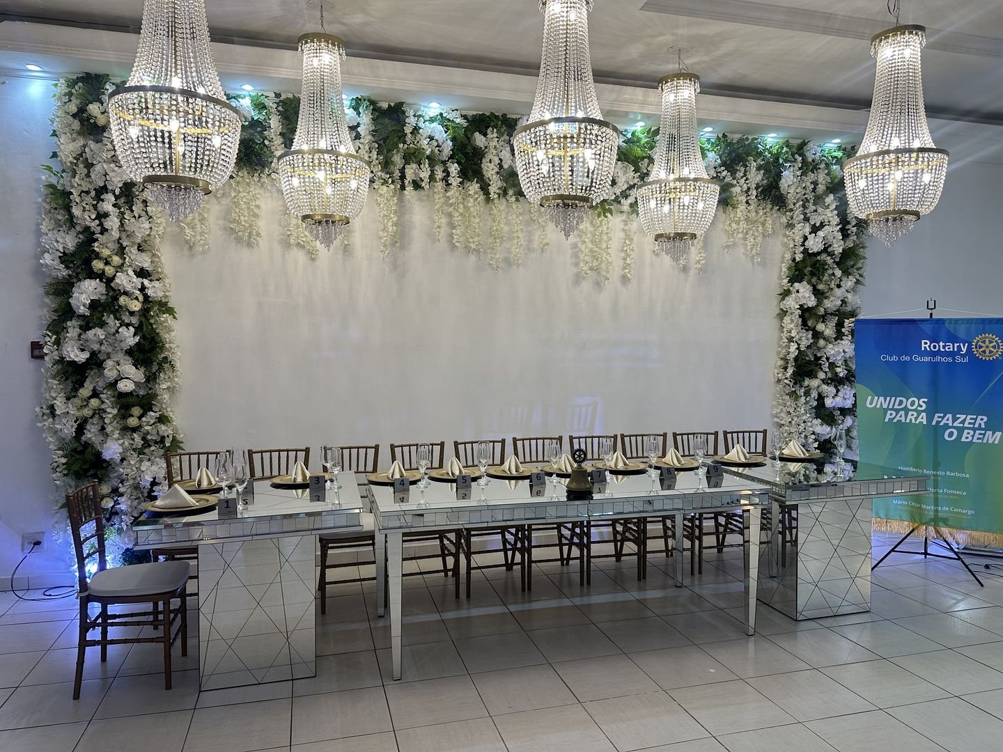 Mesa imperial montada para evento corporativo no Estrela Buffet, sob lustres de cristal e painel de flores